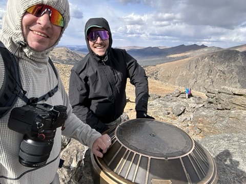 Conner and a friend at a mountain summit marker, camera in hand